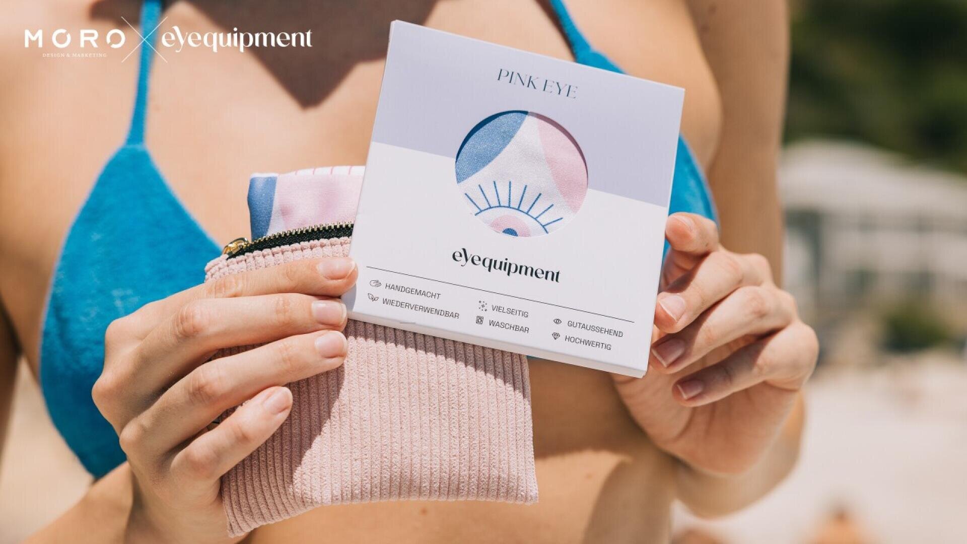 A person in a blue bikini holds a package labeled Pink Eye from the brand eYequipment, along with a pink textured pouch. The background appears to be a sunny outdoor setting, possibly a beach.