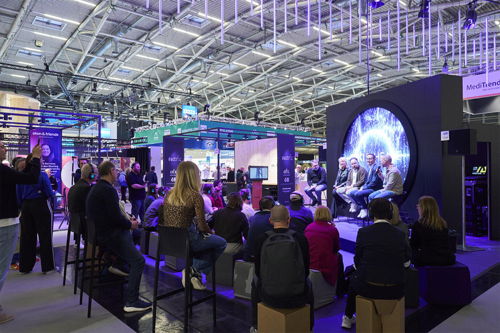 A group of people sit and listen to a panel discussion on a stage with a circular screen, inside a large, modern exhibition hall filled with booths and bright overhead lighting.