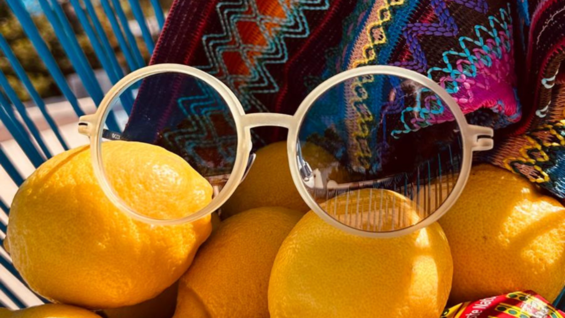 A pair of round, clear glasses rests on a pile of lemons, with a colorful woven fabric draped in the background. Blue basket spokes and sunlight create shadows and reflections on the scene.