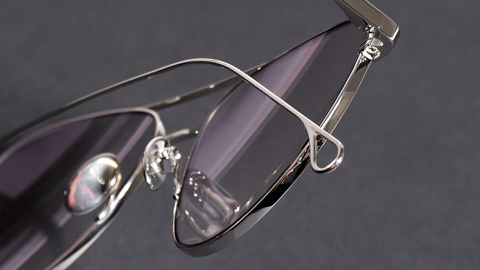 Nahaufnahme einer stylischen Sonnenbrille mit dünnem, silbernem Metallrahmen und violett getönten Gläsern vor einem dunklen Hintergrund.