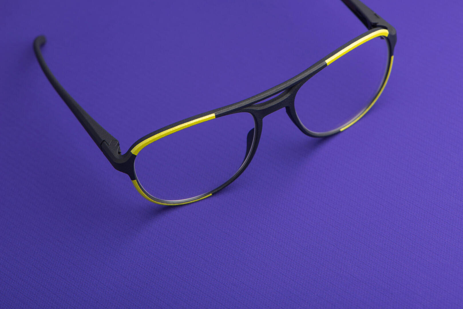 Eine schwarze Brille mit gelben Akzenten am Rahmen liegt auf einer flachen, hellvioletten, strukturierten Oberfläche.