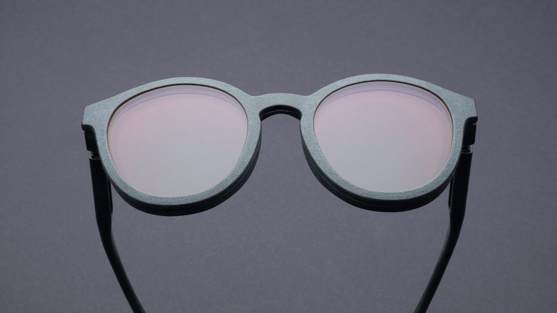 Vor einem schlichten grauen Hintergrund ist eine runde, graue Sonnenbrille mit hellrosa getönten Gläsern zu sehen.