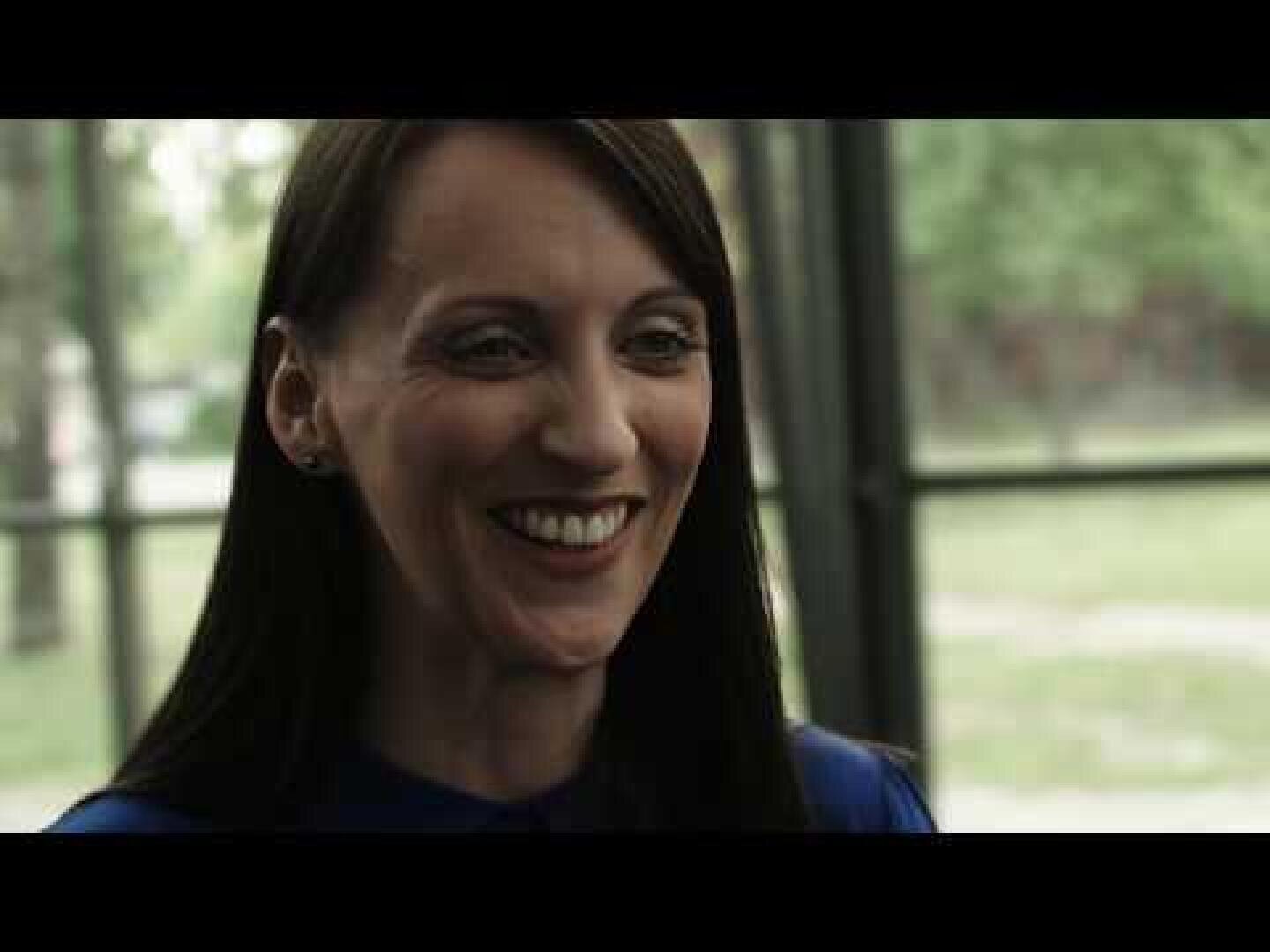 Eine Frau mit glattem dunklem Haar, die ein blaues Oberteil trägt, lächelt in einem Innenraum vor großen Fenstern mit einem verschwommenen grünen Hintergrund im Freien.