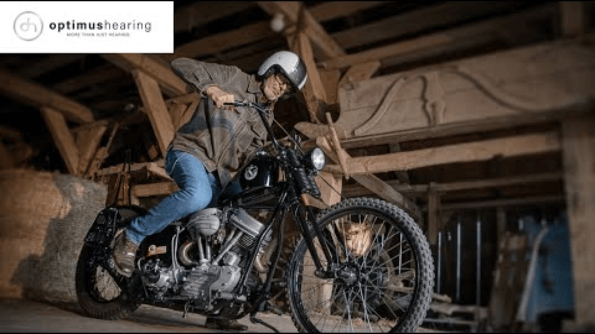 A man wearing a helmet and glasses is starting a vintage motorcycle inside a wooden barn. The logo optimushearing appears in the top left corner.