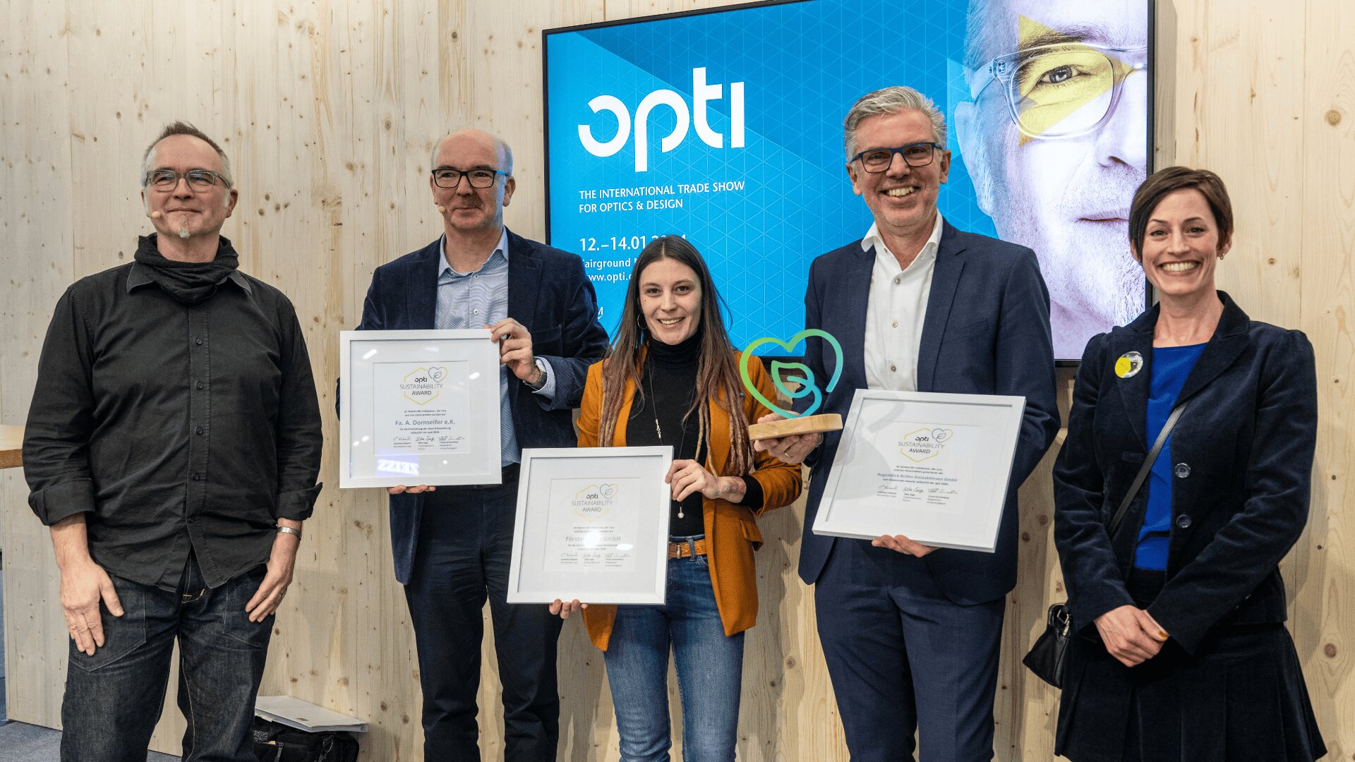 Fünf Personen stehen lächelnd in einem Raum; drei halten gerahmte Urkunden, eine hält einen herzförmigen Preis. Hinter ihnen ist ein Bildschirm mit Informationen über eine Optik- und Designmesse zu sehen.