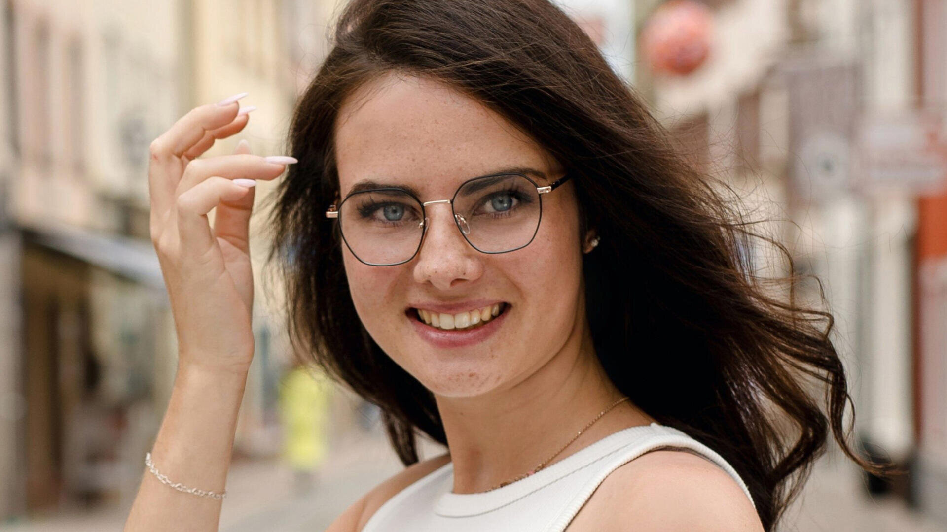 A young woman with long dark hair and glasses smiles at the camera as she stands on a city street in a sleeveless white top.