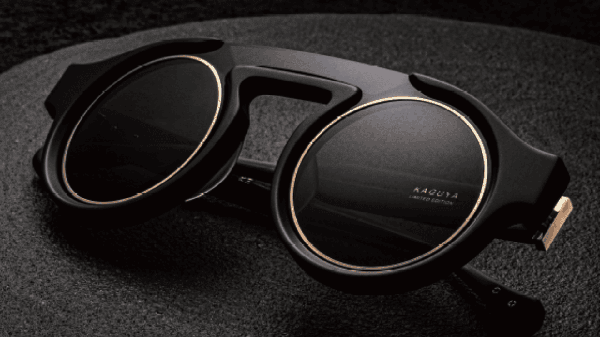 A pair of stylish black sunglasses with round lenses and gold accents rests on a dark textured surface. The frame has a bold, modern design, and KAGUYA LIMITED EDITION is visible on one lens.