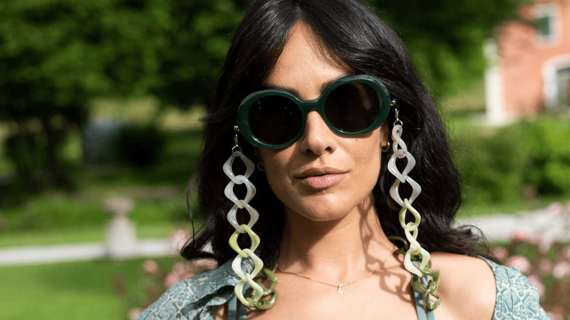 A woman with long dark hair wears large round sunglasses and bold, dangling chain-link earrings. She stands outdoors with greenery and a building in the blurred background.