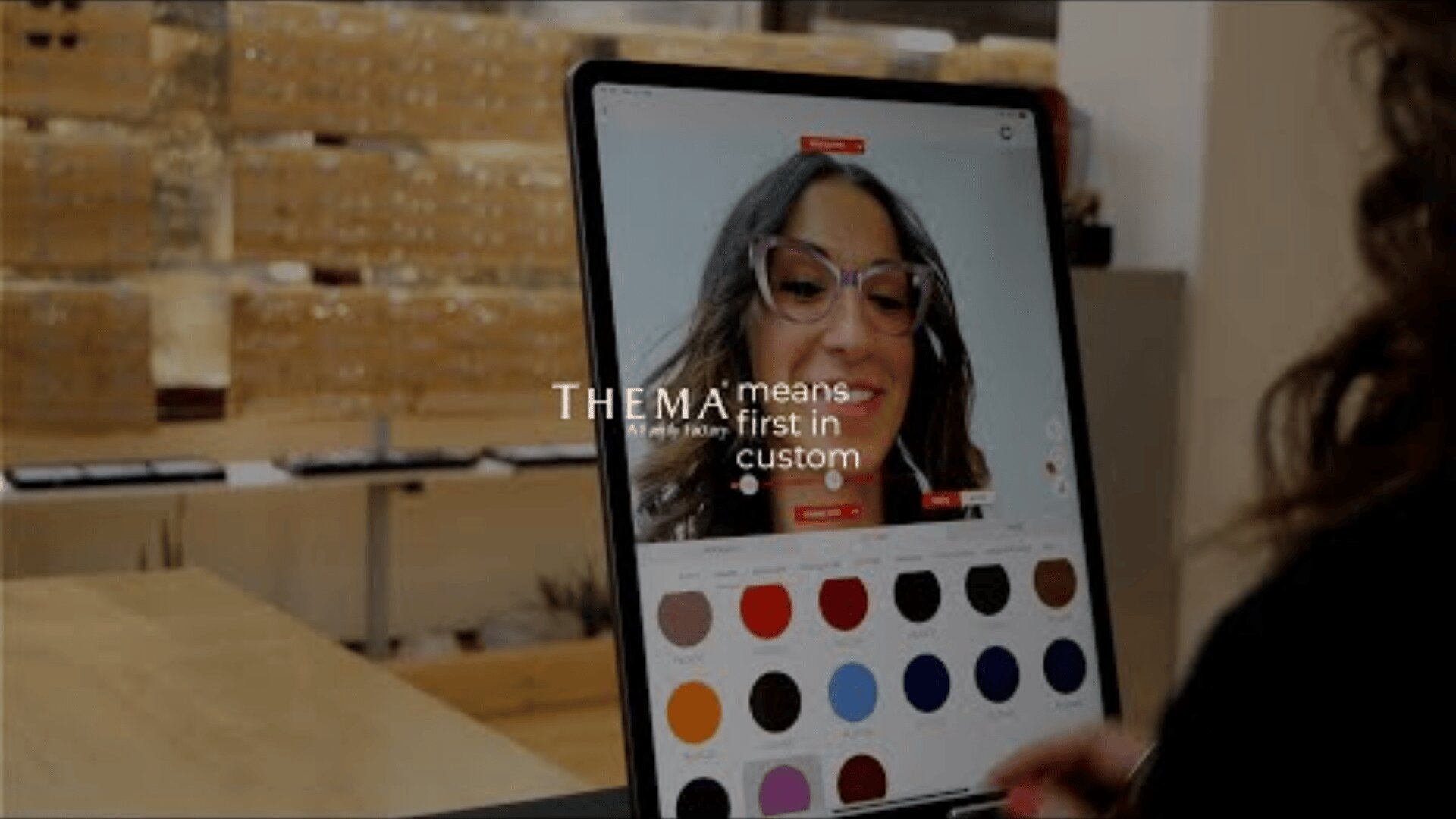 A person uses a virtual try-on tool for glasses on a computer screen, with various frame colors displayed below their face. Text on the screen reads, THEMA means first in custom.