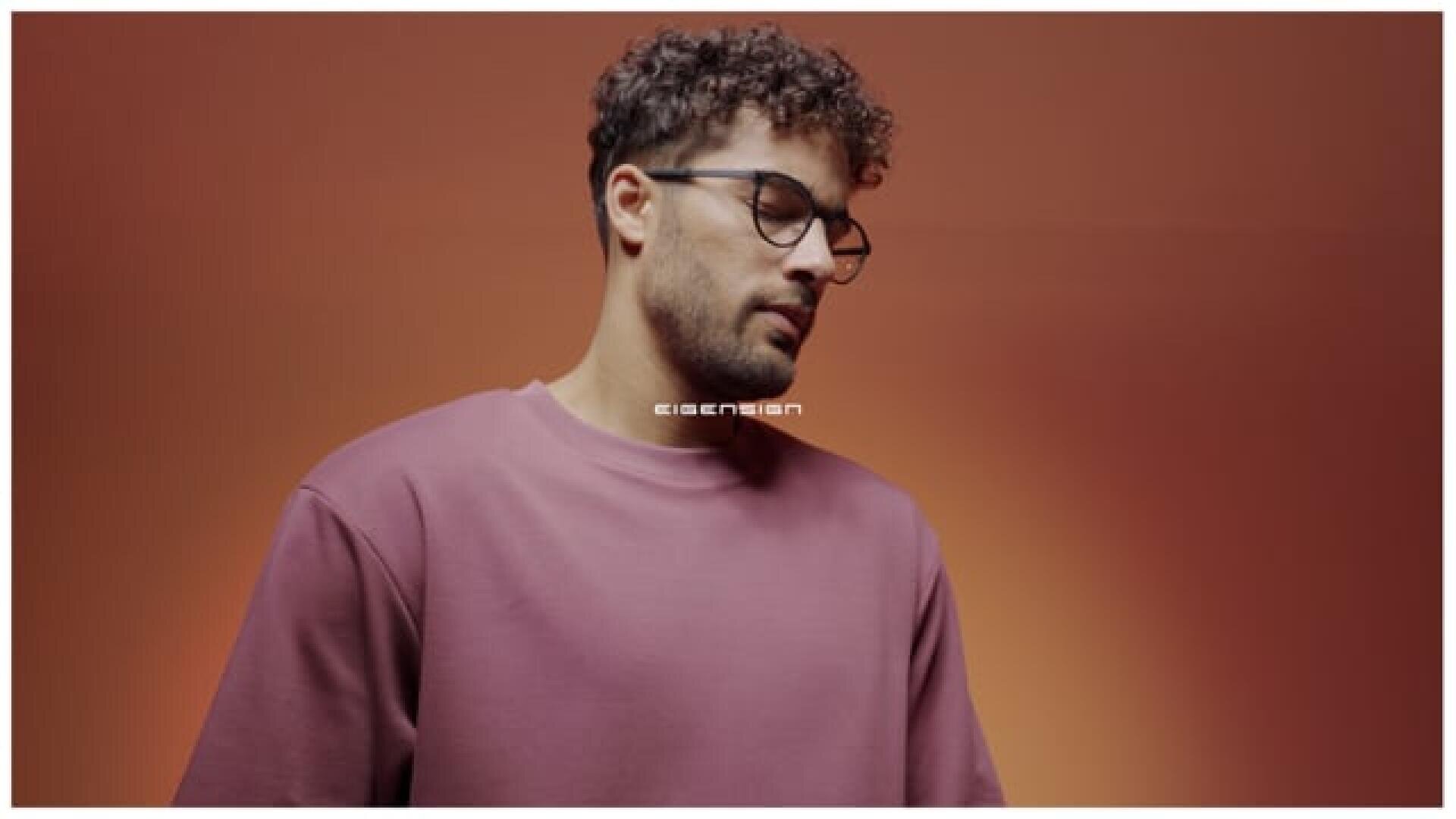 Ein Mann mit kurzen lockigen Haaren und Brille, der ein lilafarbenes Sweatshirt trägt, steht vor einem schlichten, warmen orangefarbenen Hintergrund und blickt mit neutraler Miene nach unten.