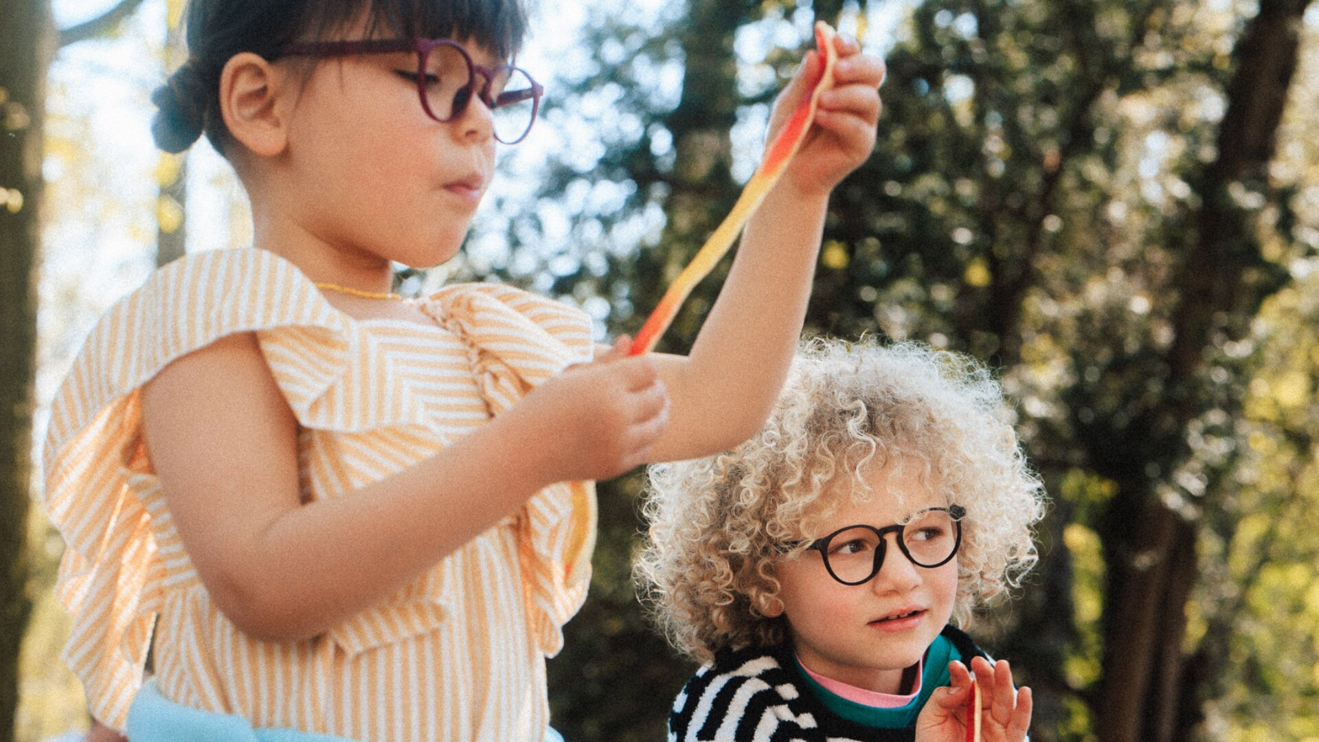 Zwei kleine Kinder mit Brillen spielen draußen. Eines hält ein buntes Band in der Hand, während das andere, mit lockigem Haar, zusieht. Das Sonnenlicht fällt durch die Bäume im Hintergrund.