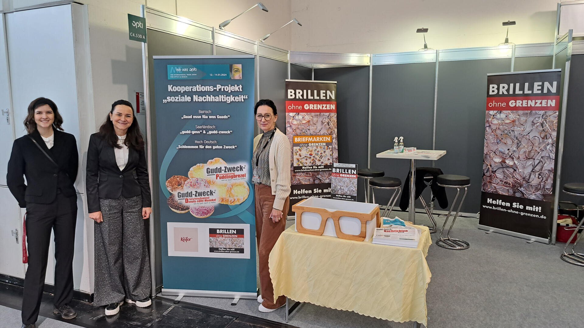 Drei Frauen stehen an einem Messestand für ein soziales Nachhaltigkeitsprojekt und Brillen ohne Grenzen. Am Stand sind Banner, Informationsmaterialien und ein Tisch mit Brillengestellen zu sehen.