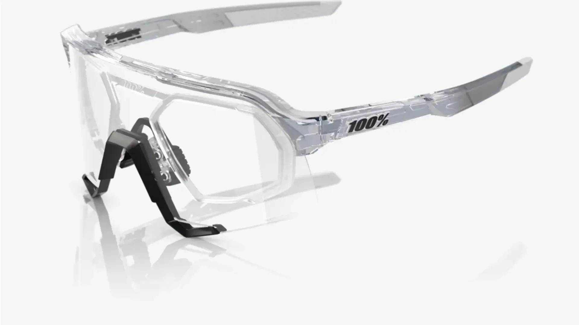 Klare Sportschutzbrille mit transparentem Rahmen und schwarzem Nasenstück, mit dem 100%-Logo an der Seite, das auf einer weißen reflektierenden Oberfläche angezeigt wird.