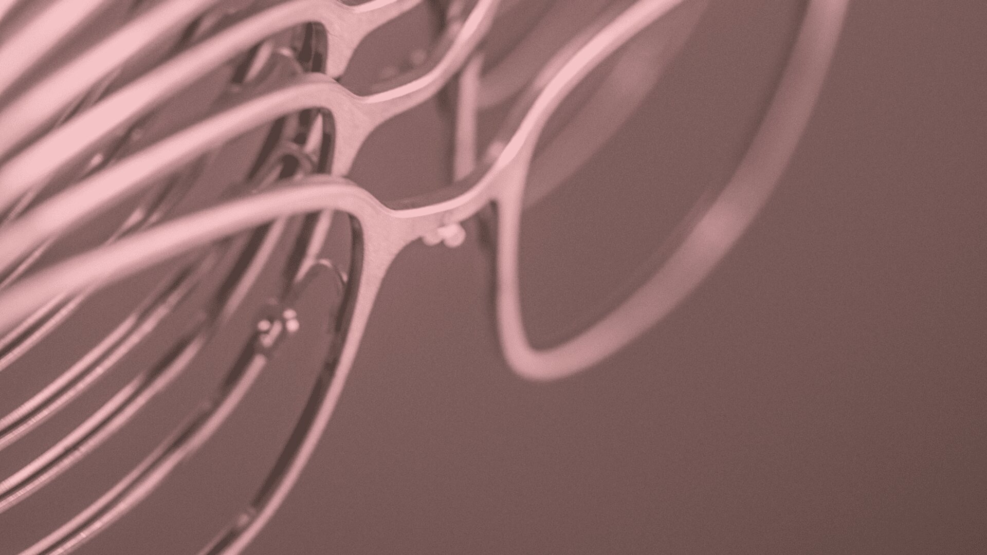A close-up view of several pairs of eyeglasses stacked together in a diagonal arrangement, with a soft pink filter over the image.