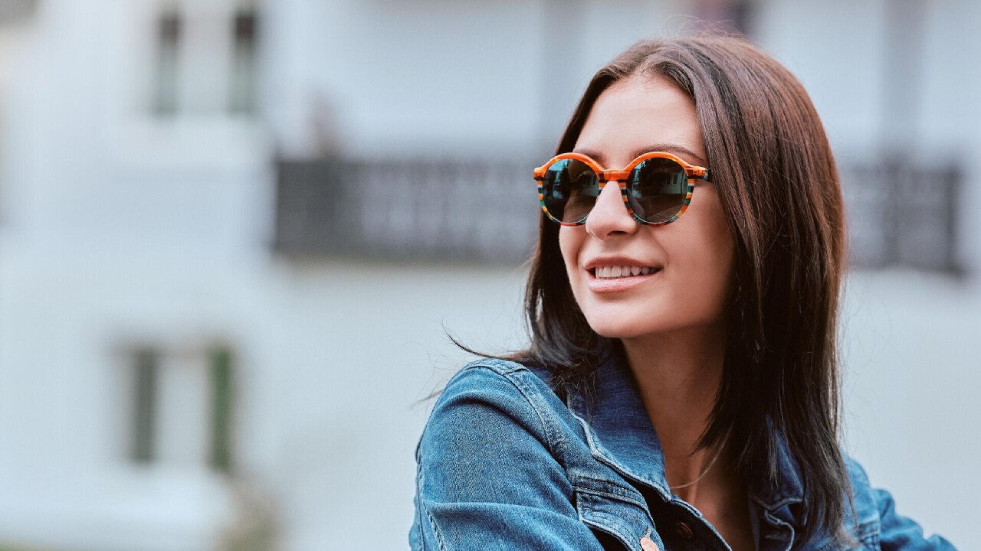 A woman with long dark hair wearing sunglasses and a denim jacket smiles while looking to the side, with a blurred building in the background.