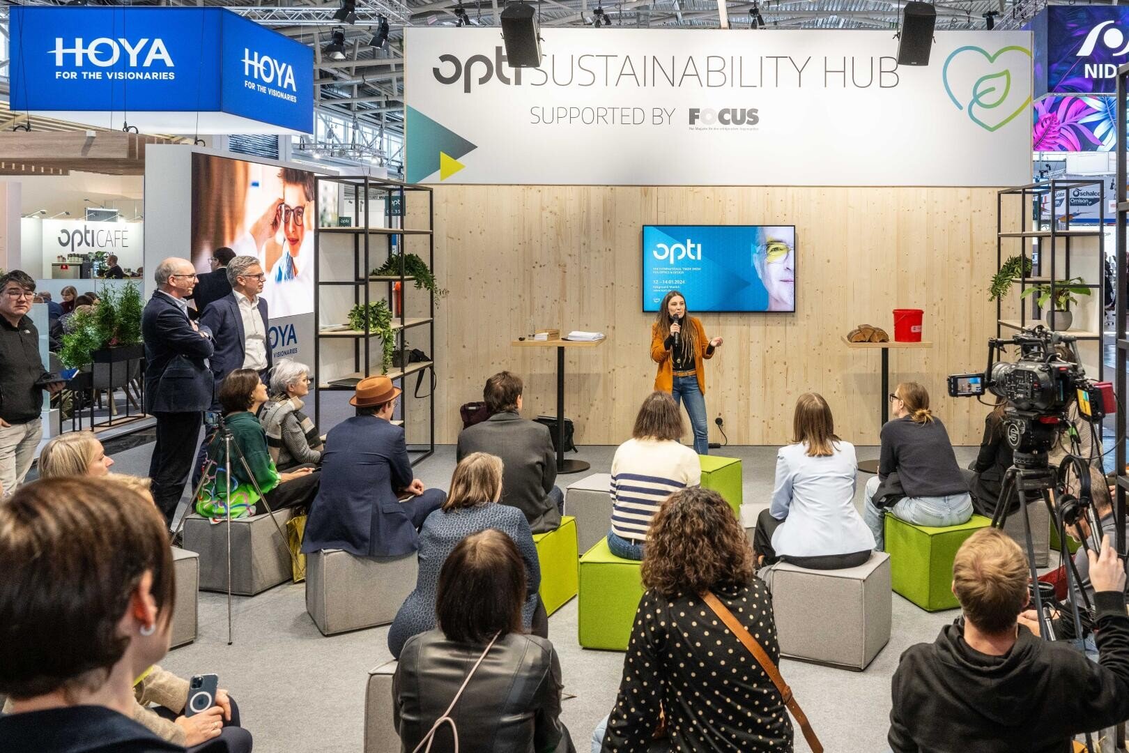 Eine Frau spricht auf der Bühne des opti Sustainability Hub. Das Publikum sitzt auf Würfelhockern. Der Veranstaltungsort ist hell und modern, mit Bildschirmen und Bannern, die das Event-Branding und die Logos der Sponsoren zeigen.