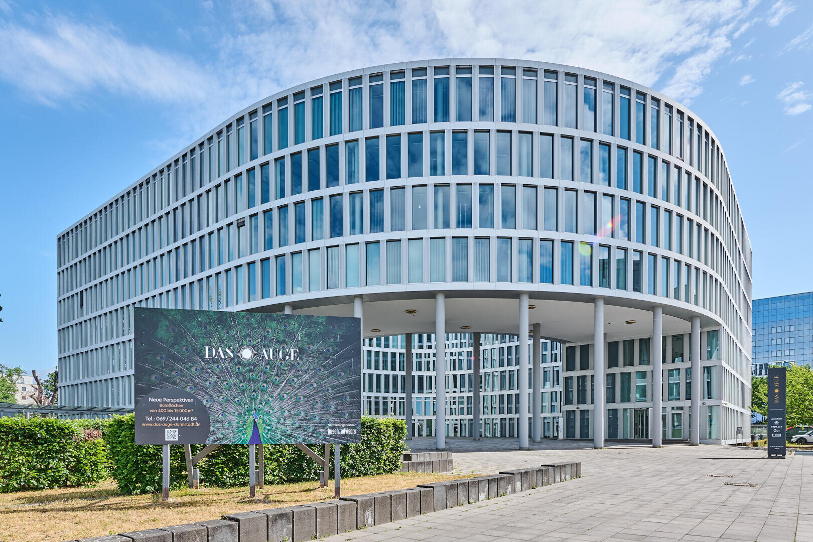 A modern, curved glass office building with a white frame stands behind a large billboard and green plants under a blue sky with scattered clouds.