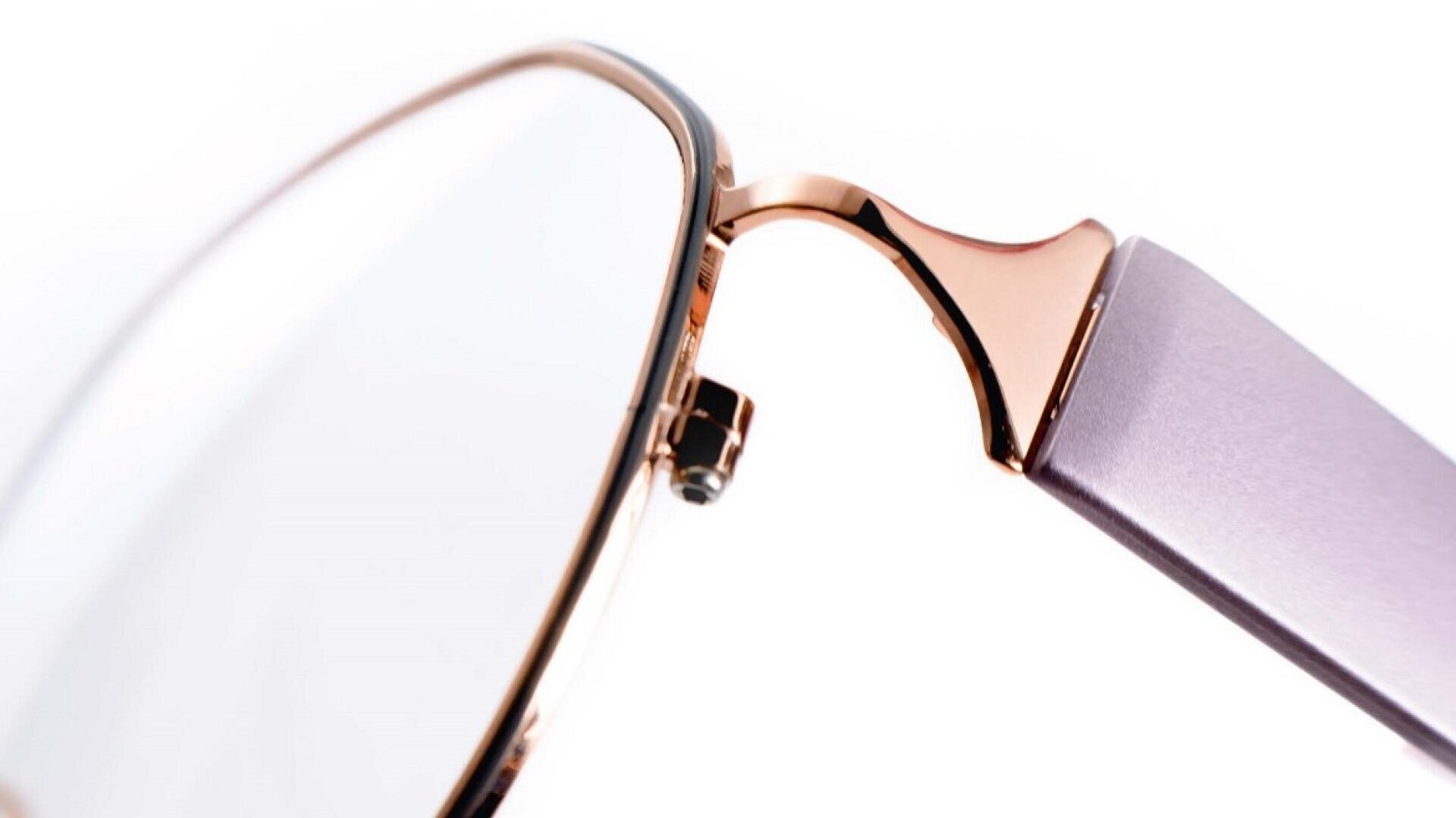 Nahaufnahme des Scharniers und des Rahmens einer roségoldenen Metallbrille mit lavendelfarbenen Bügeln vor einem weißen Hintergrund.