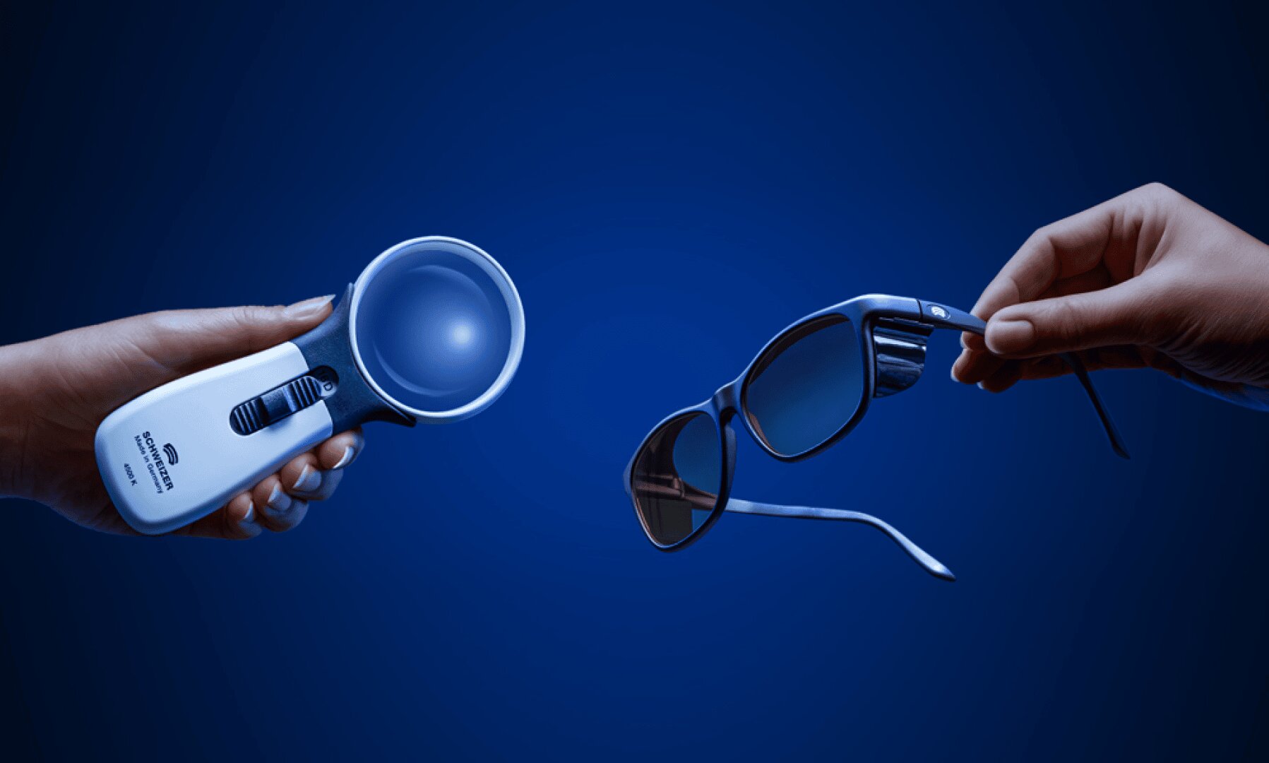Die linke Hand hält ein Vergrößerungsglas mit Licht, die rechte Hand hält eine Sonnenbrille vor blauem Hintergrund.