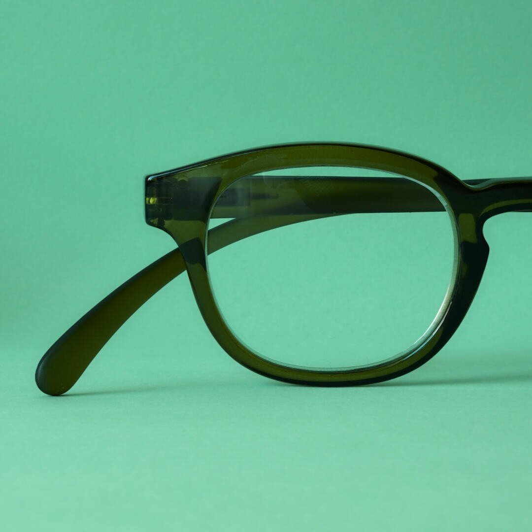 Eine Nahaufnahme einer dunkelgrünen Brille mit dickem Rahmen, die sich vor einem passenden hellgrünen Hintergrund befindet. Nur die rechte Seite der Brille ist sichtbar.