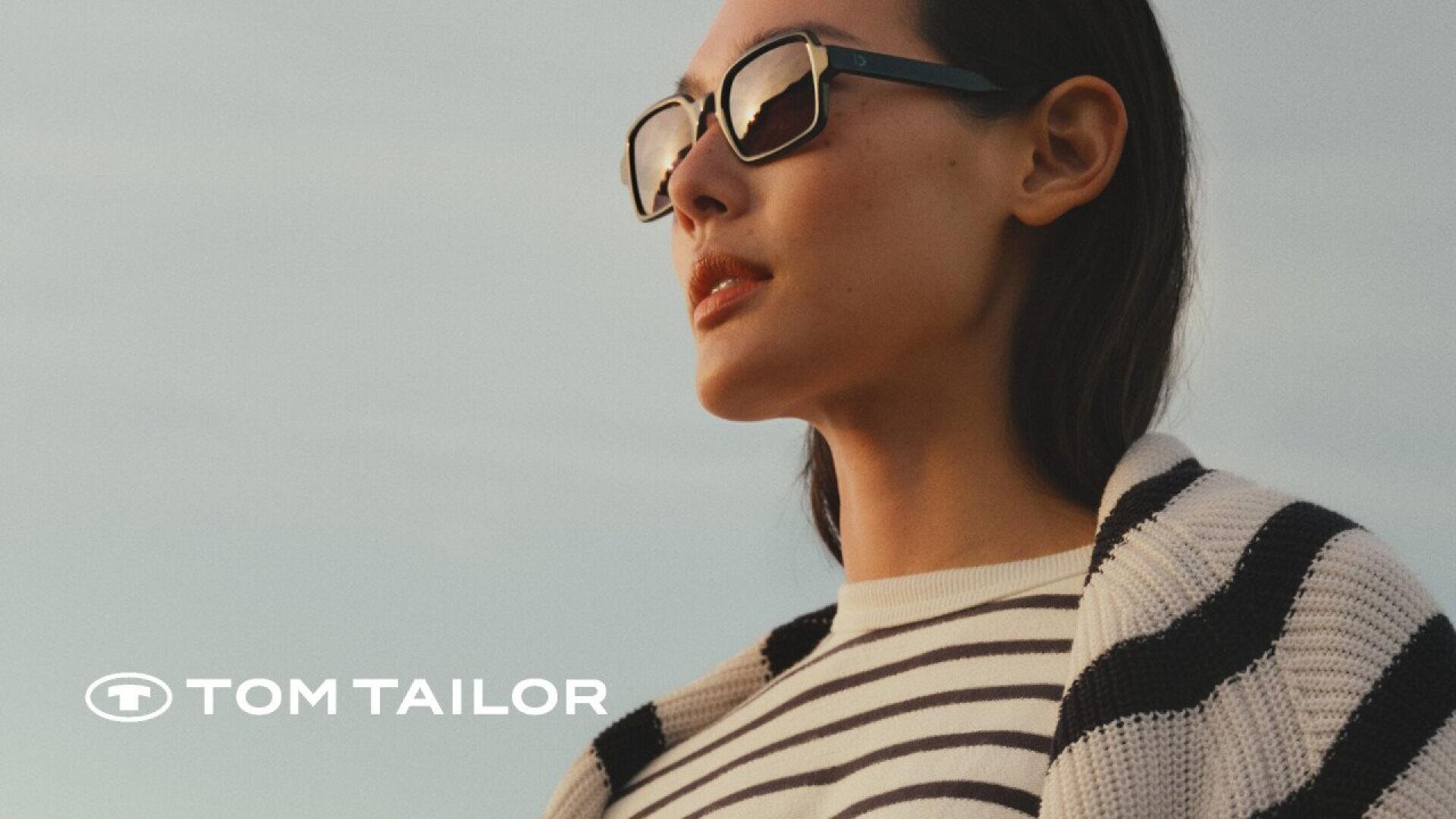 A woman wearing sunglasses and a striped shirt with a striped sweater draped over her shoulders looks into the distance. The Tom Tailor logo appears in the lower left corner.