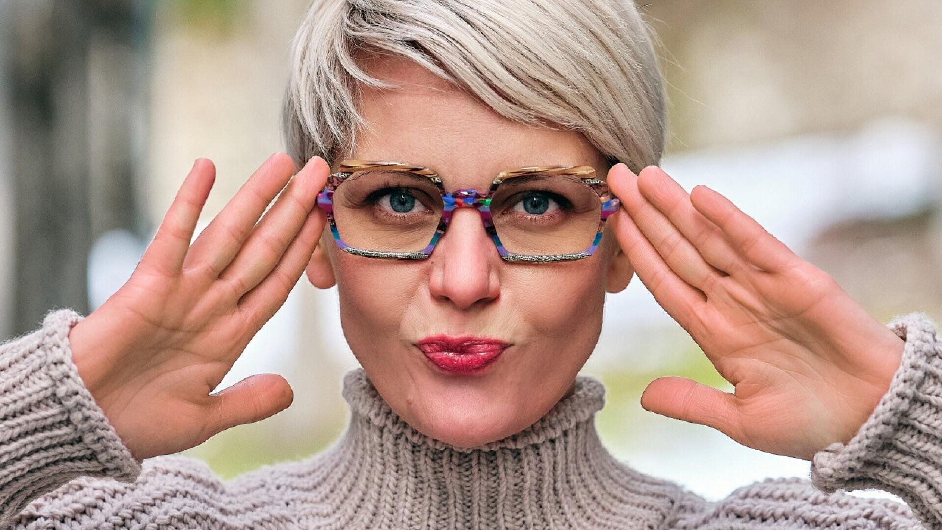 A woman with short blonde hair and colorful glasses poses playfully with her hands raised near her face, wearing a chunky knit sweater and pursing her lips. The background is softly blurred.