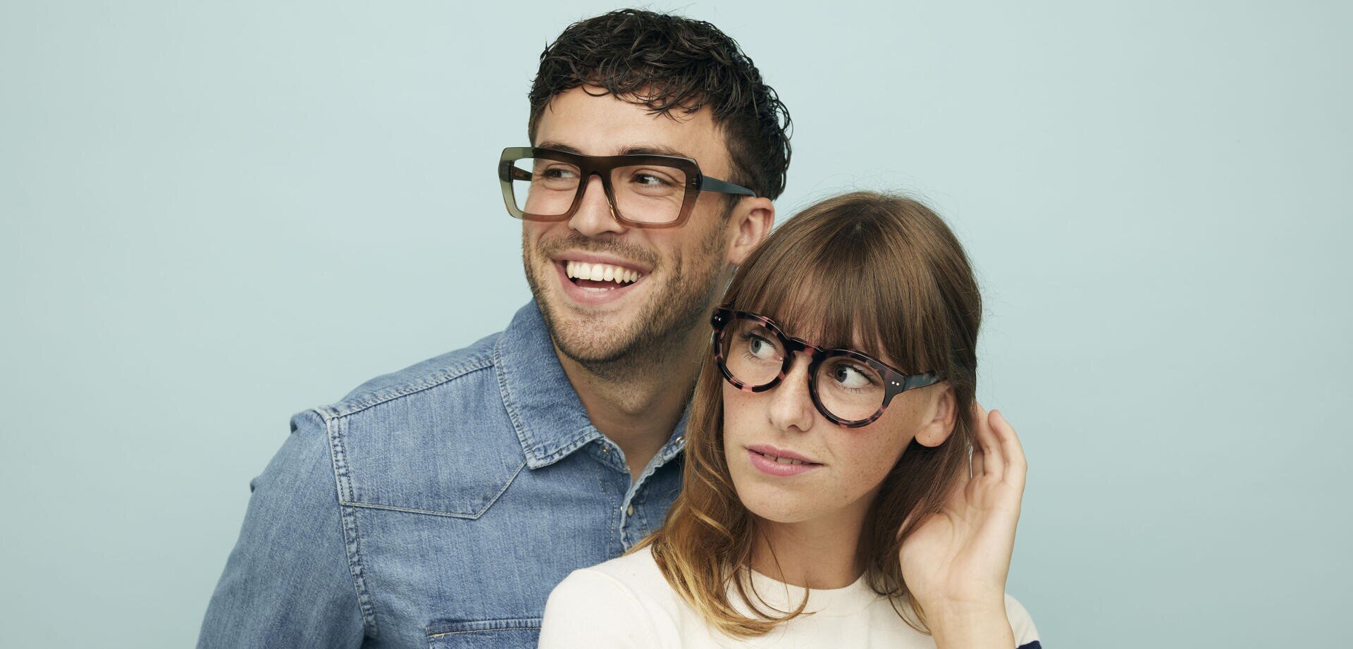 Ein Mann in einem Jeanshemd und eine Frau in einem weißen Oberteil, beide mit großen Brillengläsern, stehen vor einem hellblauen Hintergrund. Der Mann lächelt und schaut nach links, während die Frau zu ihm aufschaut und ihr Haar berührt.