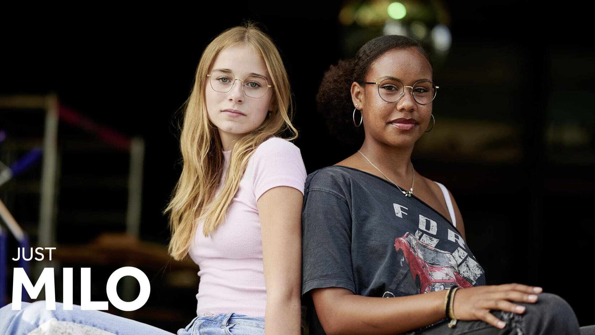 Zwei junge Frauen sitzen Rücken an Rücken, beide tragen Brillen und legere Kleidung und schauen selbstbewusst in die Kamera. In der linken unteren Ecke erscheint der Text "JUST MILO".