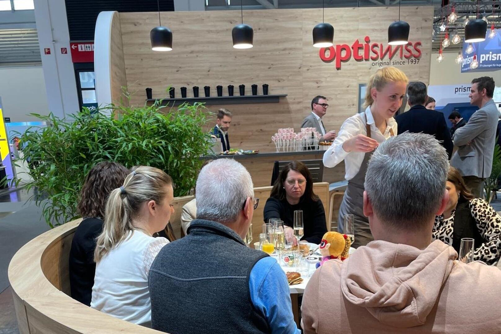 Eine Gruppe von Personen sitzt um einen Tisch mit Speisen und Getränken, während ein Kellner in der Nähe steht. Dahinter befindet sich ein hölzerner Stand mit dem optiswiss-Logo und Regalen mit Bechern, in einer Messe- oder Ausstellungsumgebung.