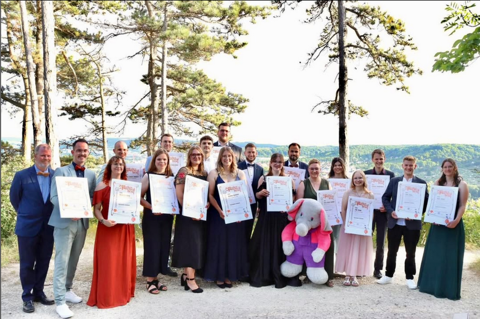 A group of people dressed formally stand outdoors holding certificates, smiling at the camera. Trees and a scenic landscape are in the background. A person in a large, pink-and-gray elephant costume stands with them.