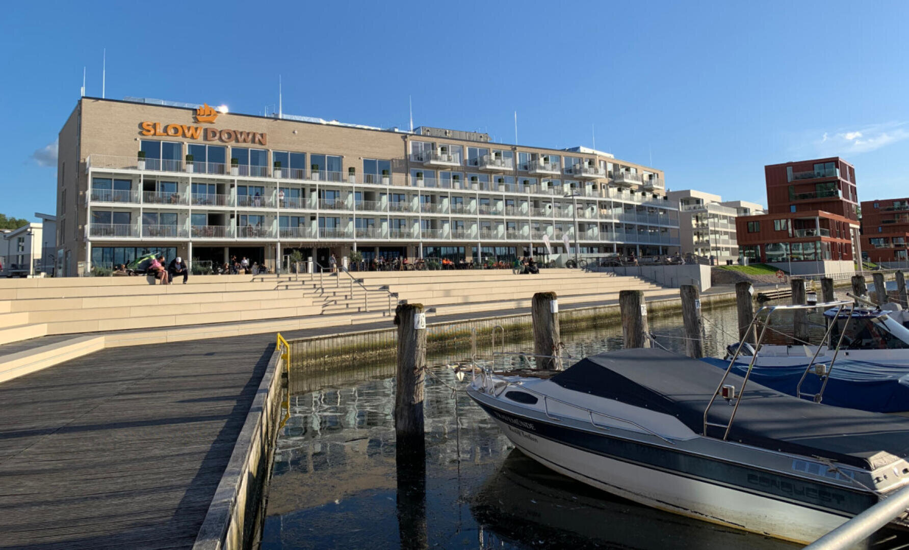 Ein modernes Hotel am Wasser mit Glasbalkonen und der Aufschrift „SLOW DOWN“ liegt gegenüber einem Jachthafen mit vertäuten Booten, Holzpfosten und breiten Steintreppen unter einem klaren blauen Himmel.