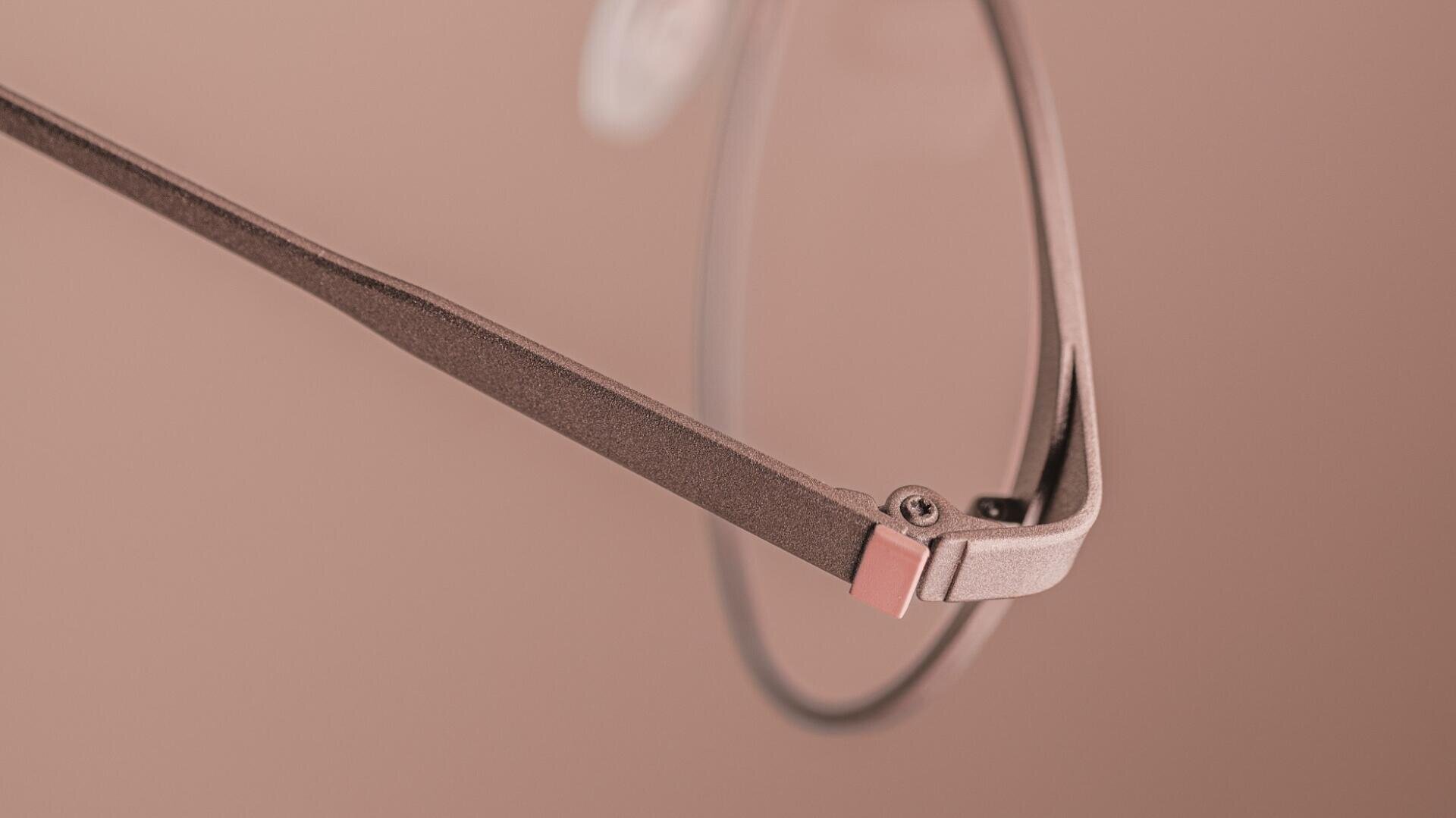 Close-up of the arm and hinge of a pair of eyeglasses with a metallic, rose-colored finish on a matching pink background. The focus is on the details of the frame and screw.