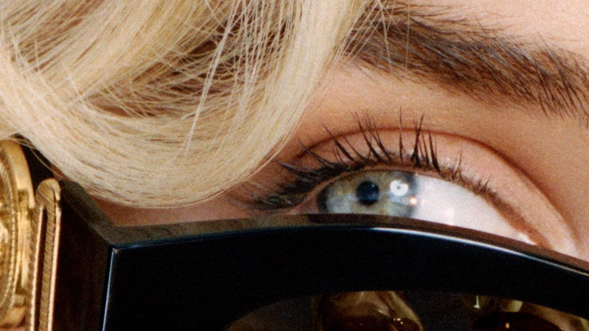 Nahaufnahme eines blauen Auges, umrahmt von langen Wimpern, hellblondem Haar und einer großen schwarzen Sonnenbrille mit einem goldenen Scharnier.