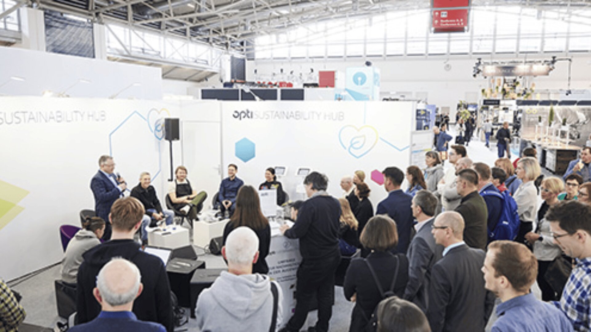 Eine bunt gemischte Gruppe von Menschen nimmt an einer Podiumsdiskussion im Rahmen einer Nachhaltigkeitsveranstaltung in einer großen, gut beleuchteten Ausstellungshalle teil. Die Diskussionsteilnehmer sitzen auf einer Bühne, während das Publikum aufmerksam zuhört.