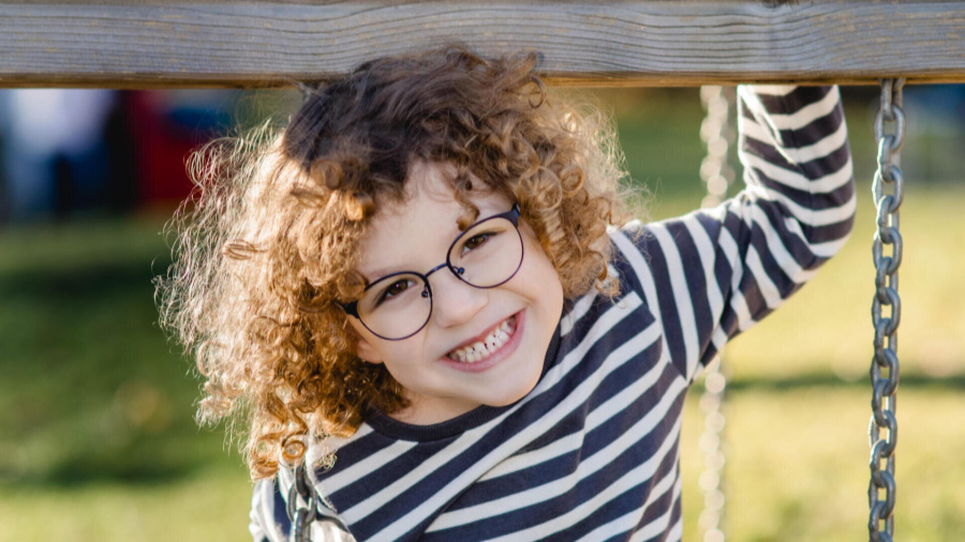A smiling child with curly hair and glasses hangs from a wooden pole on a swing, wears a black and white striped shirt and has a meadow in the background.