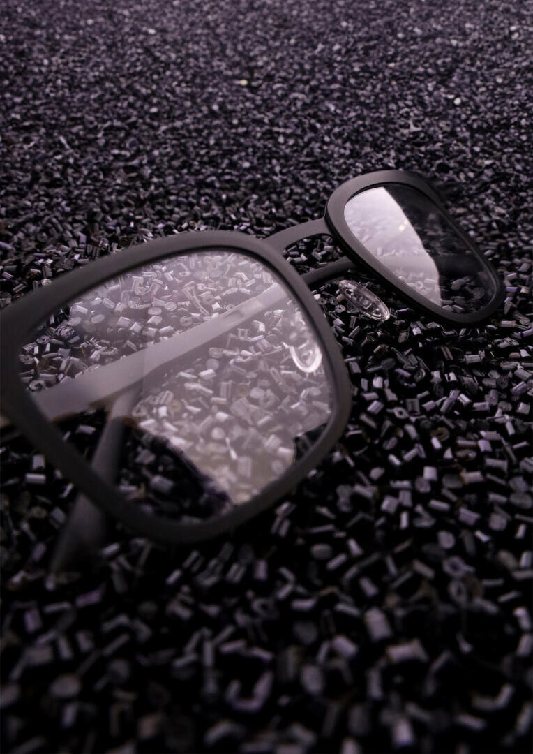 A pair of black-framed eyeglasses lies on a surface covered with many small, shiny black plastic pellets, reflecting light in the lenses.