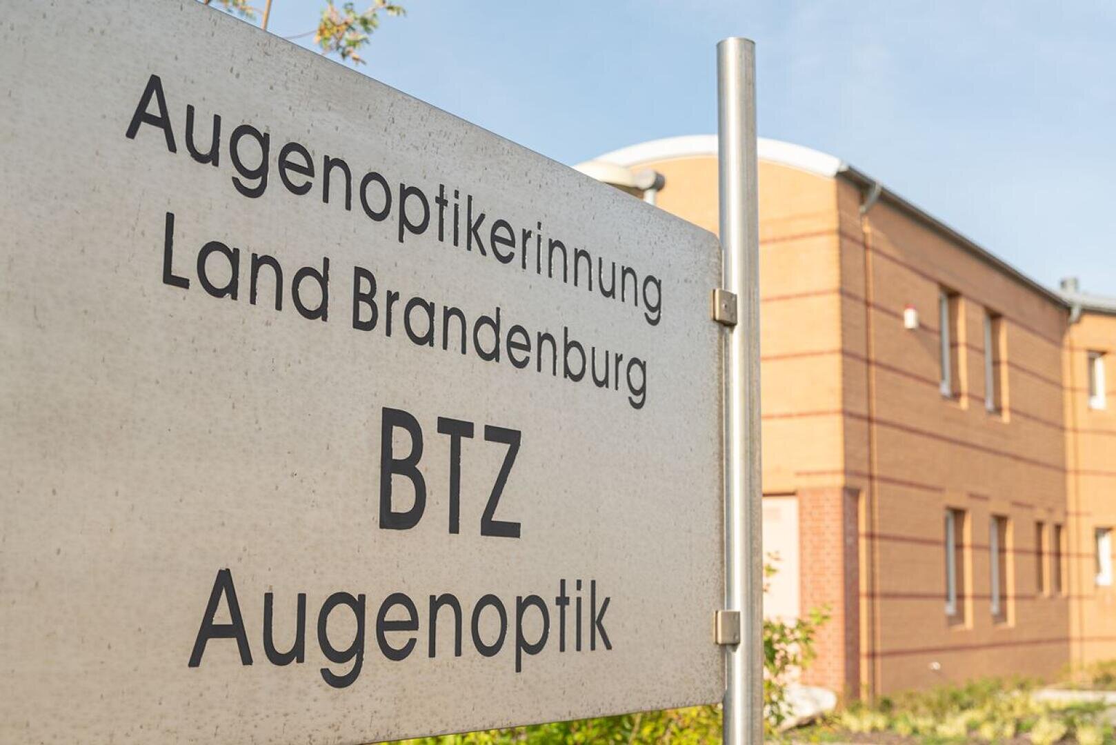 Ein Schild mit der Aufschrift Augenoptikerinnung Land Brandenburg BTZ Augenoptik steht an einem sonnigen Tag vor einem modernen Backsteinbau.