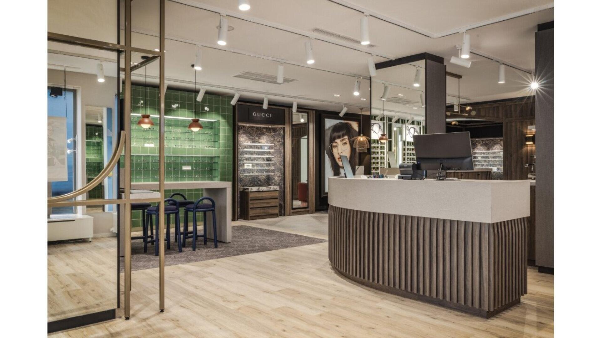 Modern eyewear store interior with wooden floors, a curved reception desk, display shelves, green accent wall, and seating area. Glasses and sunglasses are displayed on illuminated shelves, creating a stylish and welcoming atmosphere.