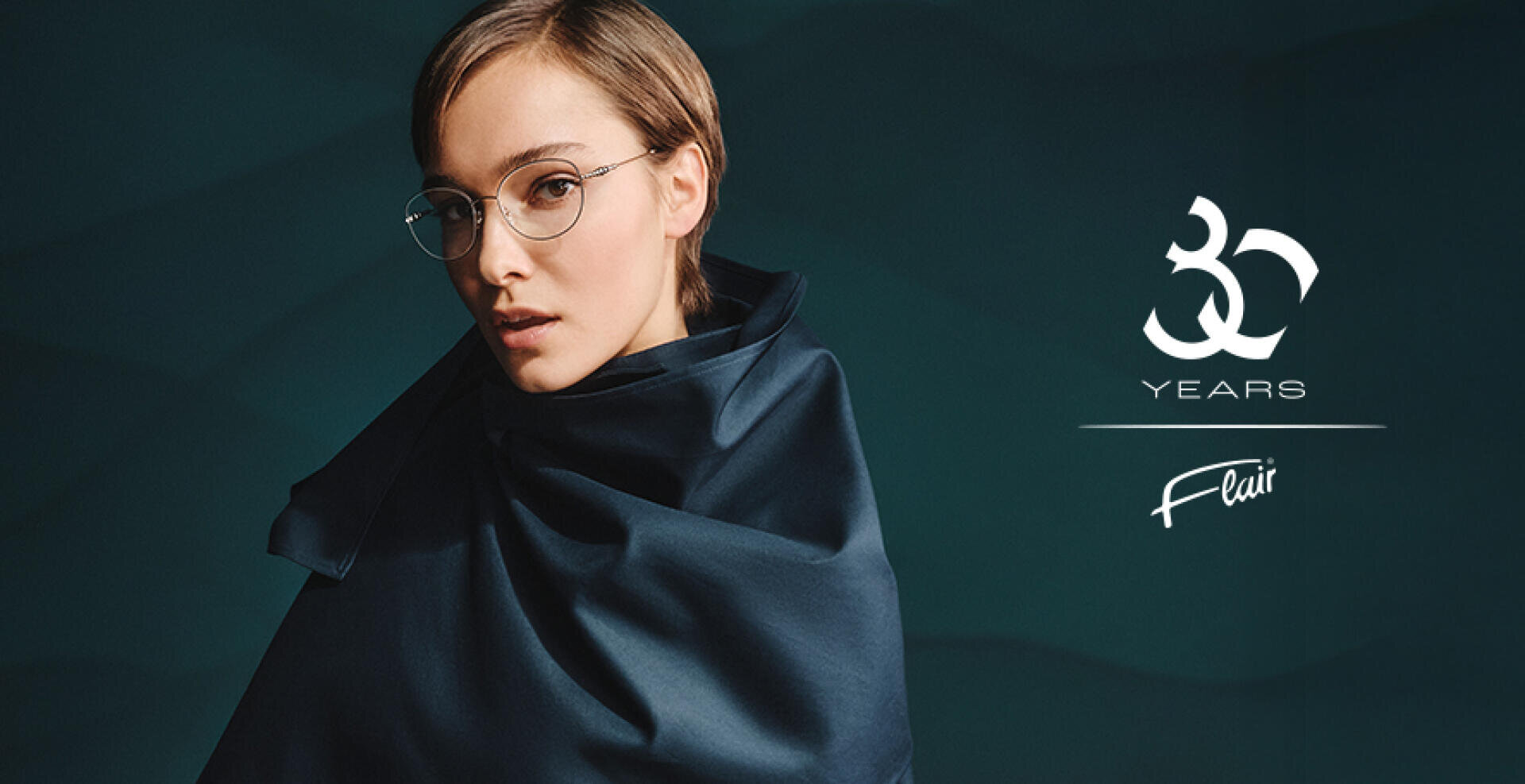 Eine Person mit einer runden Brille und einem dunklen, seidigen Schal steht vor einem dunklen, strukturierten Hintergrund. Der Text lautet 30 Jahre Flair, ein Jubiläum.