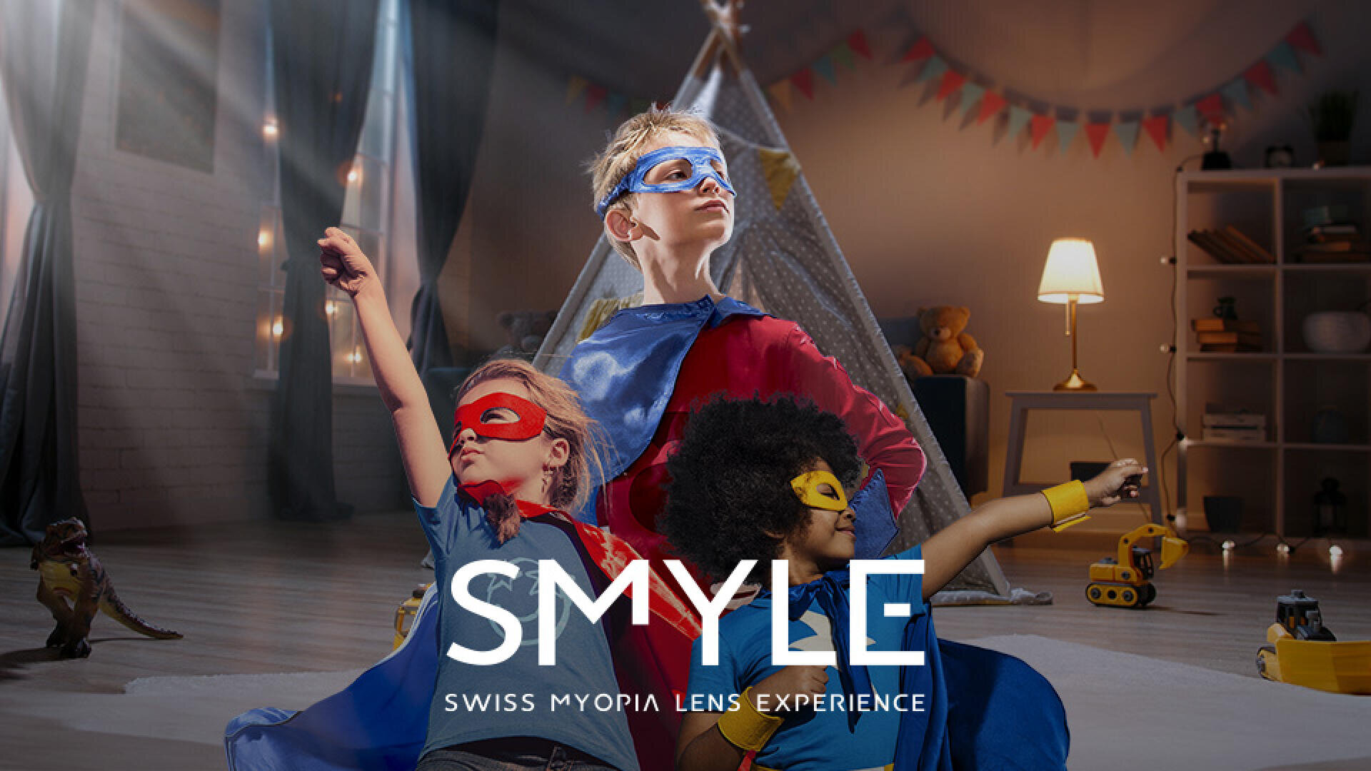 Drei Kinder in bunten Superheldenkostümen posieren selbstbewusst in einem gemütlich eingerichteten Wohnzimmer mit Spielzeug auf dem Boden. Der Text SMYLE SWISS MYOPIA LENS EXPERIENCE wird unten angezeigt.
