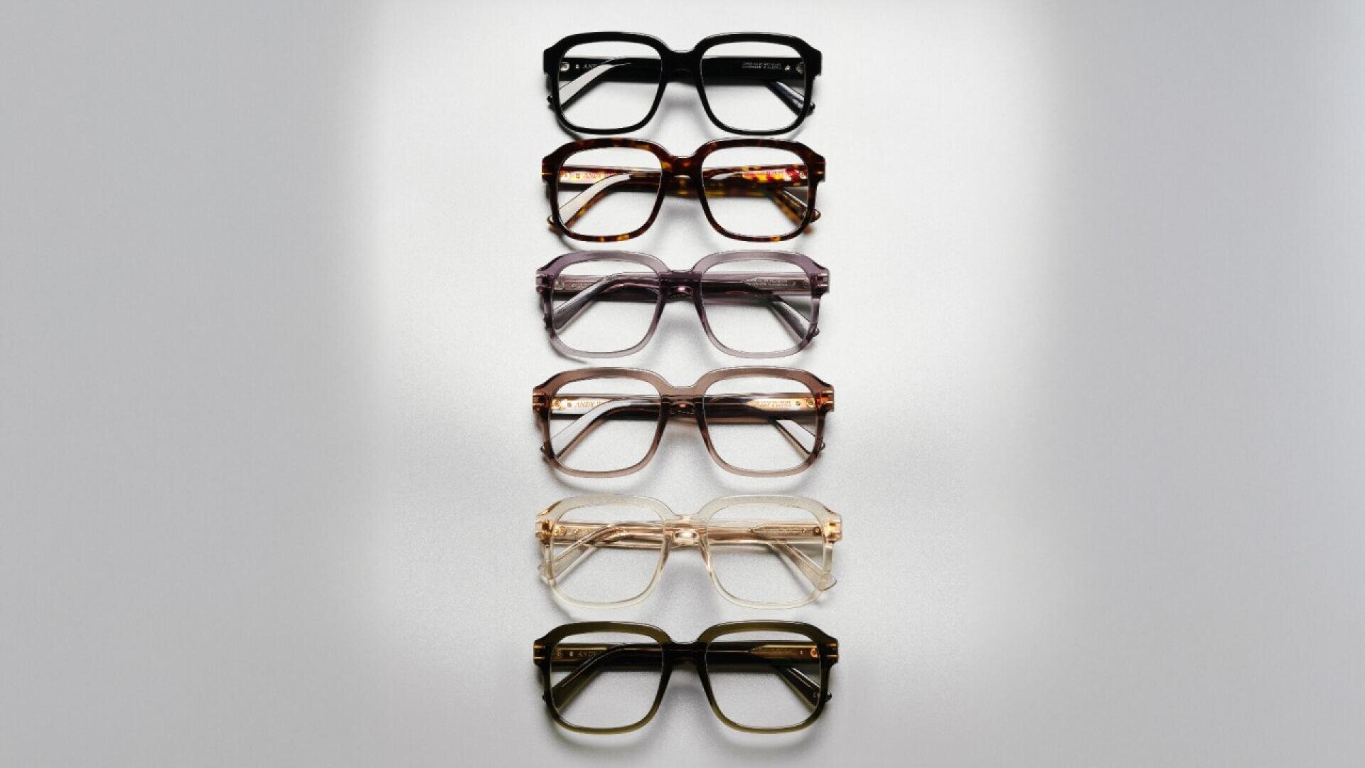 A top-down view of six pairs of eyeglasses arranged vertically in a row, each with a different frame color, ranging from dark to light shades. The background is a smooth, light gray surface.