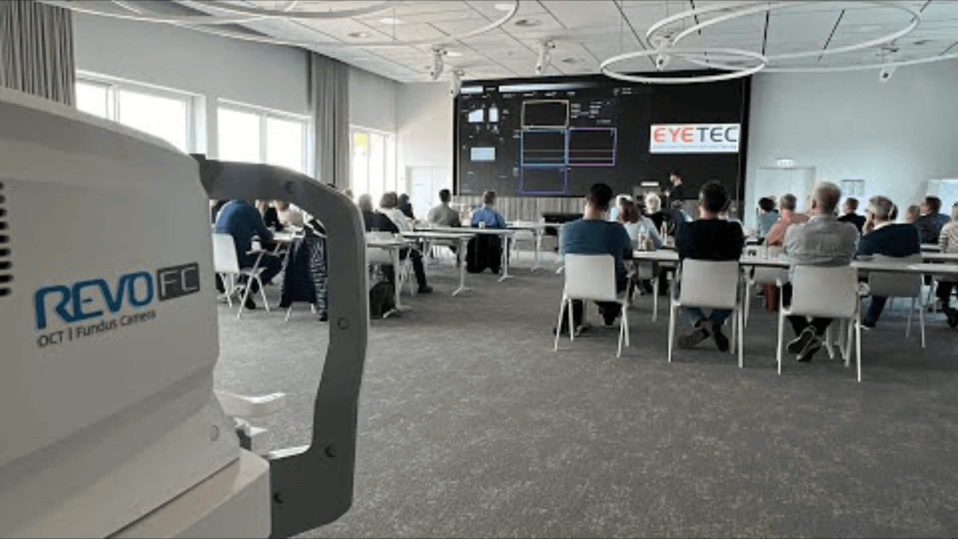 Eine Gruppe von Menschen sitzt an Tischen in einem modernen Konferenzraum und verfolgt eine Präsentation. Auf einem großen Bildschirm ist das EYETEC-Logo zu sehen und im Vordergrund ist eine OCT-Funduskamera mit der Bezeichnung REVO FC zu sehen.