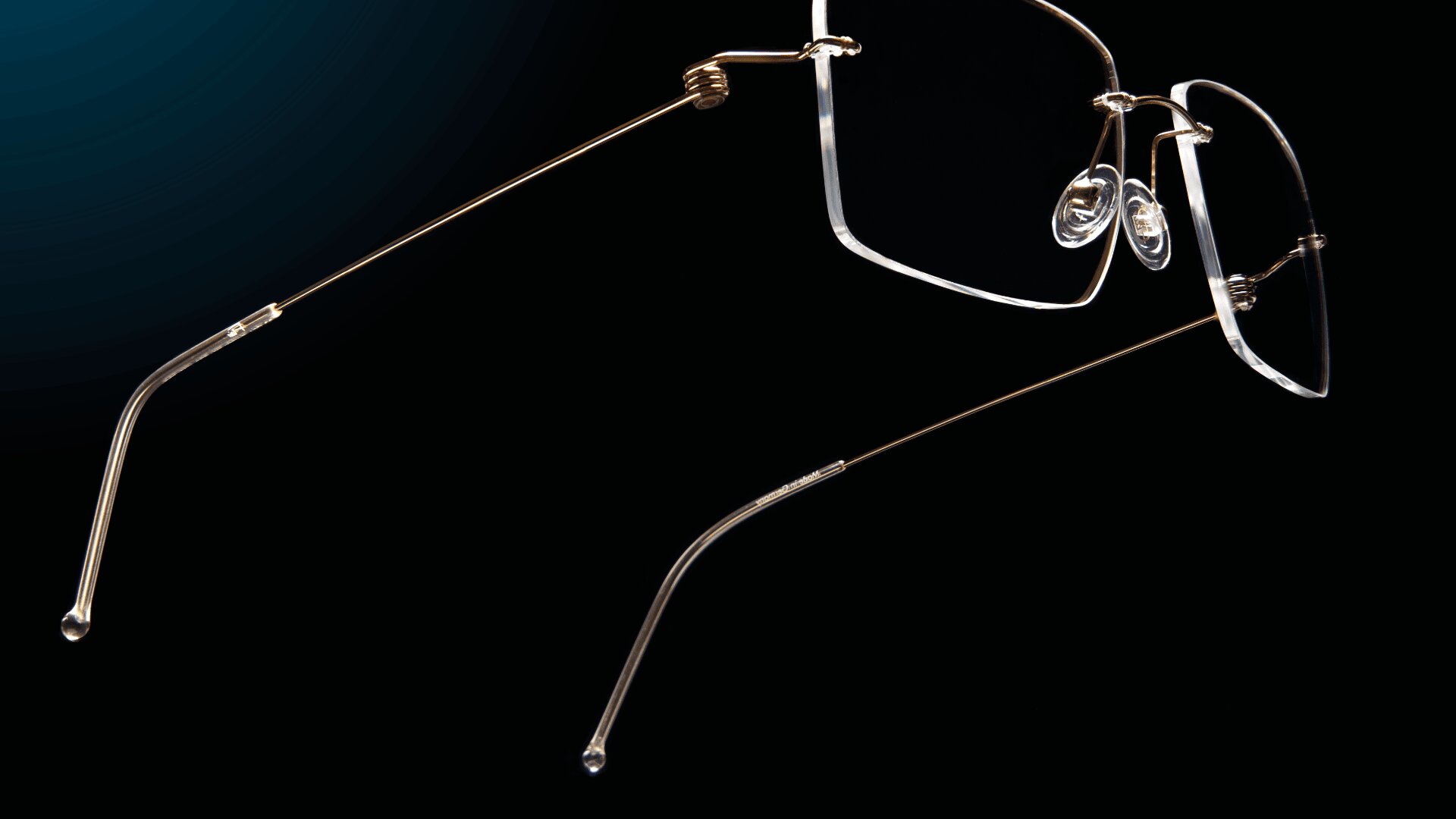 Nahaufnahme einer randlosen Brille mit dünnen goldenen Bügeln und transparenten Nasenpads vor einem schwarzen Hintergrund.