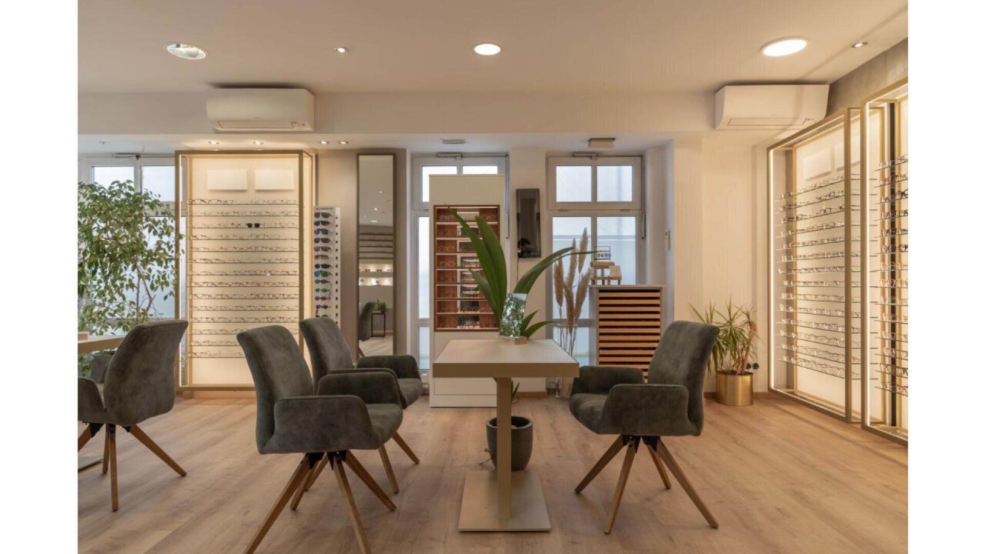 A modern eyewear store interior with display cases of eyeglasses, four gray velvet chairs around a table, and a tall green plant in the center. The room has wooden floors and large windows letting in natural light.