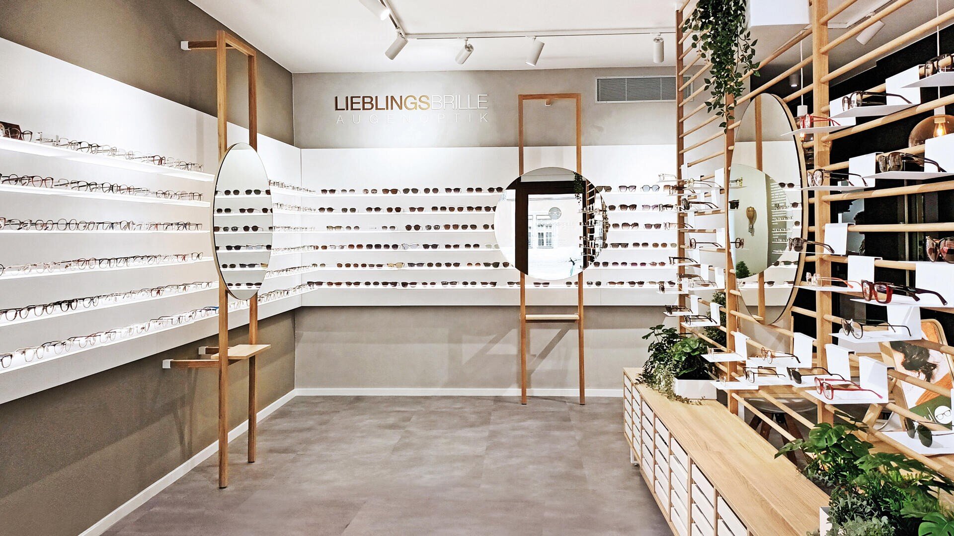 Ein modernes Brillengeschäft mit weißen Regalen, in denen viele Brillen ausgestellt sind, runden Spiegeln auf Holzständern, Pflanzen und einem minimalistischen Design. An der Rückwand befindet sich das Schild LIEBLINGSBRILLE.