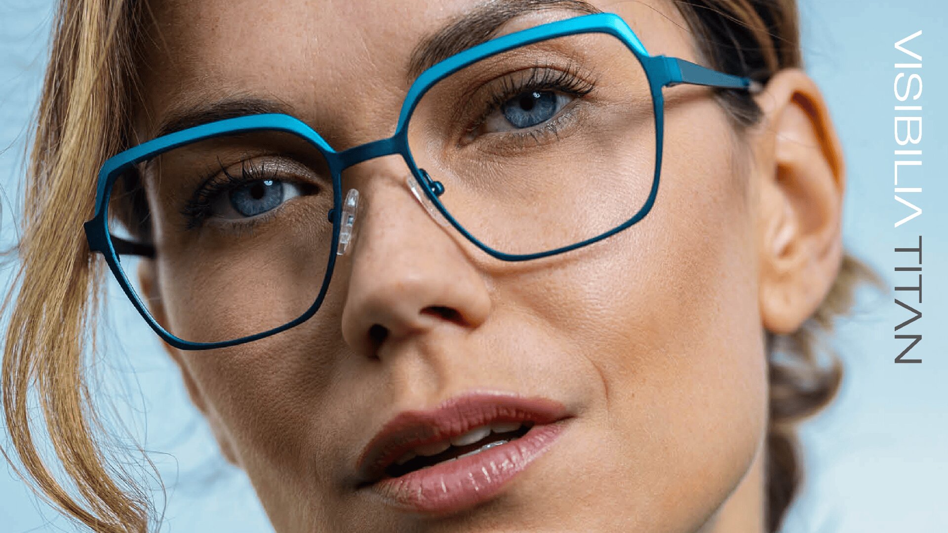 Nahaufnahme einer Frau mit einer großen, geometrischen blauen Brille, die direkt in die Kamera schaut. Der Hintergrund ist hellblau, und der Text VISIBILIA TITAN erscheint vertikal auf der rechten Seite.
