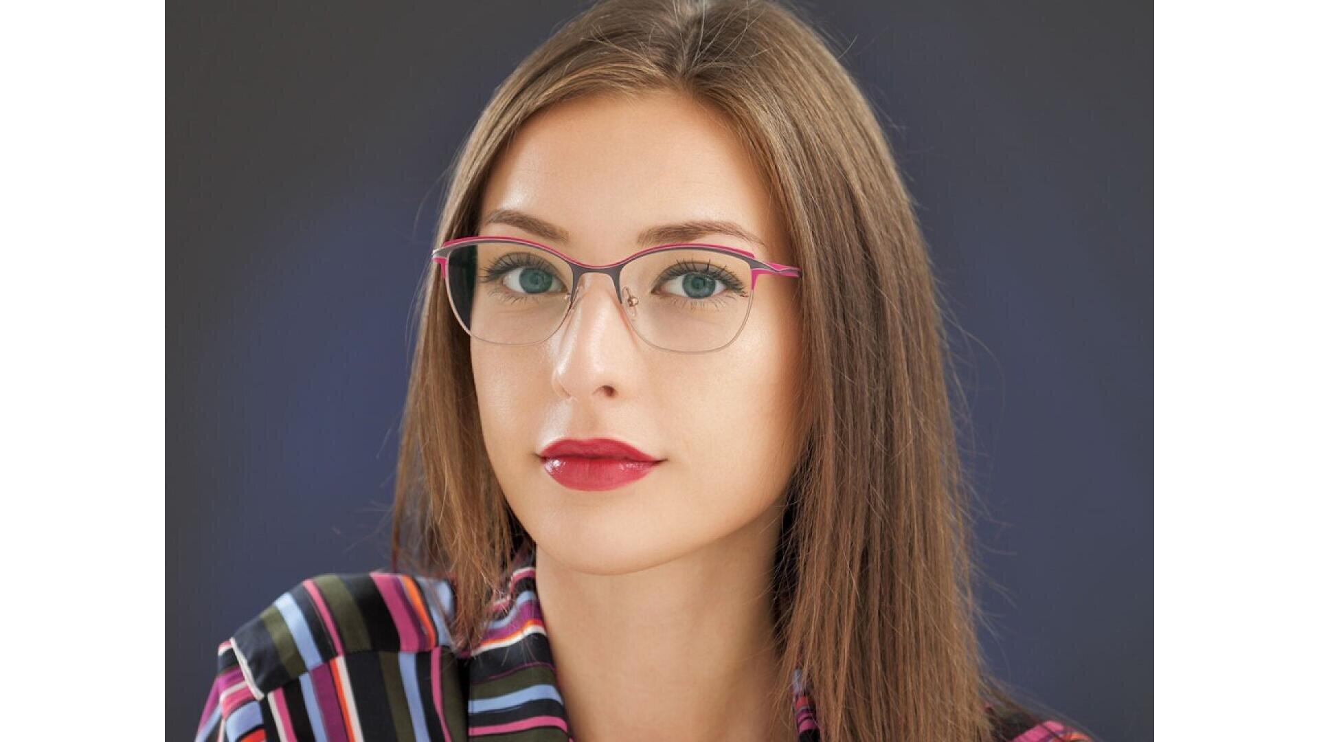 A woman with long brown hair and glasses looks at the camera. She is wearing a striped shirt and has red lipstick. The background is dark and plain.