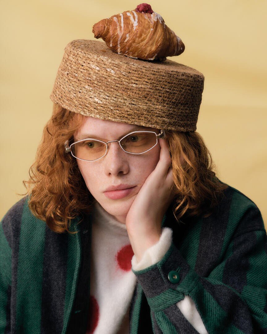 A person with curly red hair and glasses rests their face on their hand. They wear a green striped jacket and have a straw hat topped with a croissant on their head, against a pale yellow background.