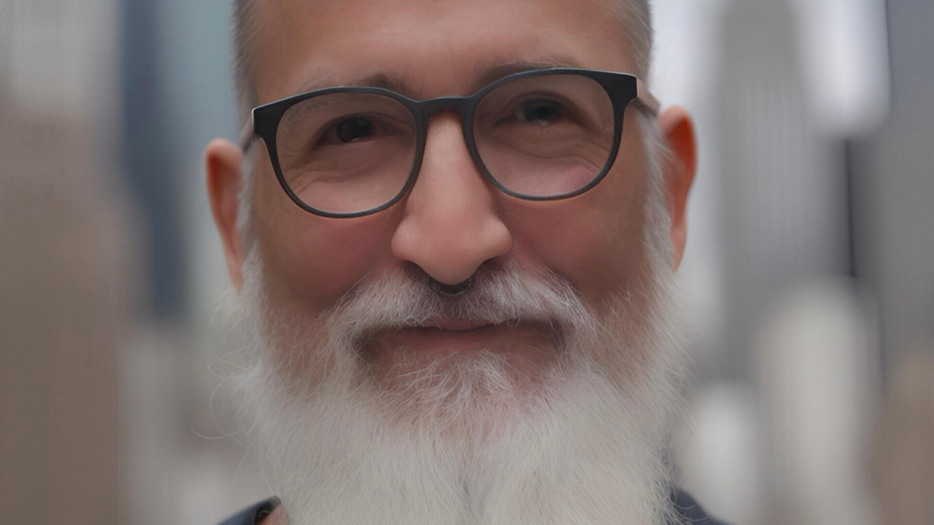Ein lächelnder älterer Mann mit weißem Bart und Brille steht im Freien, im Hintergrund sind verschwommene Stadtgebäude zu sehen.