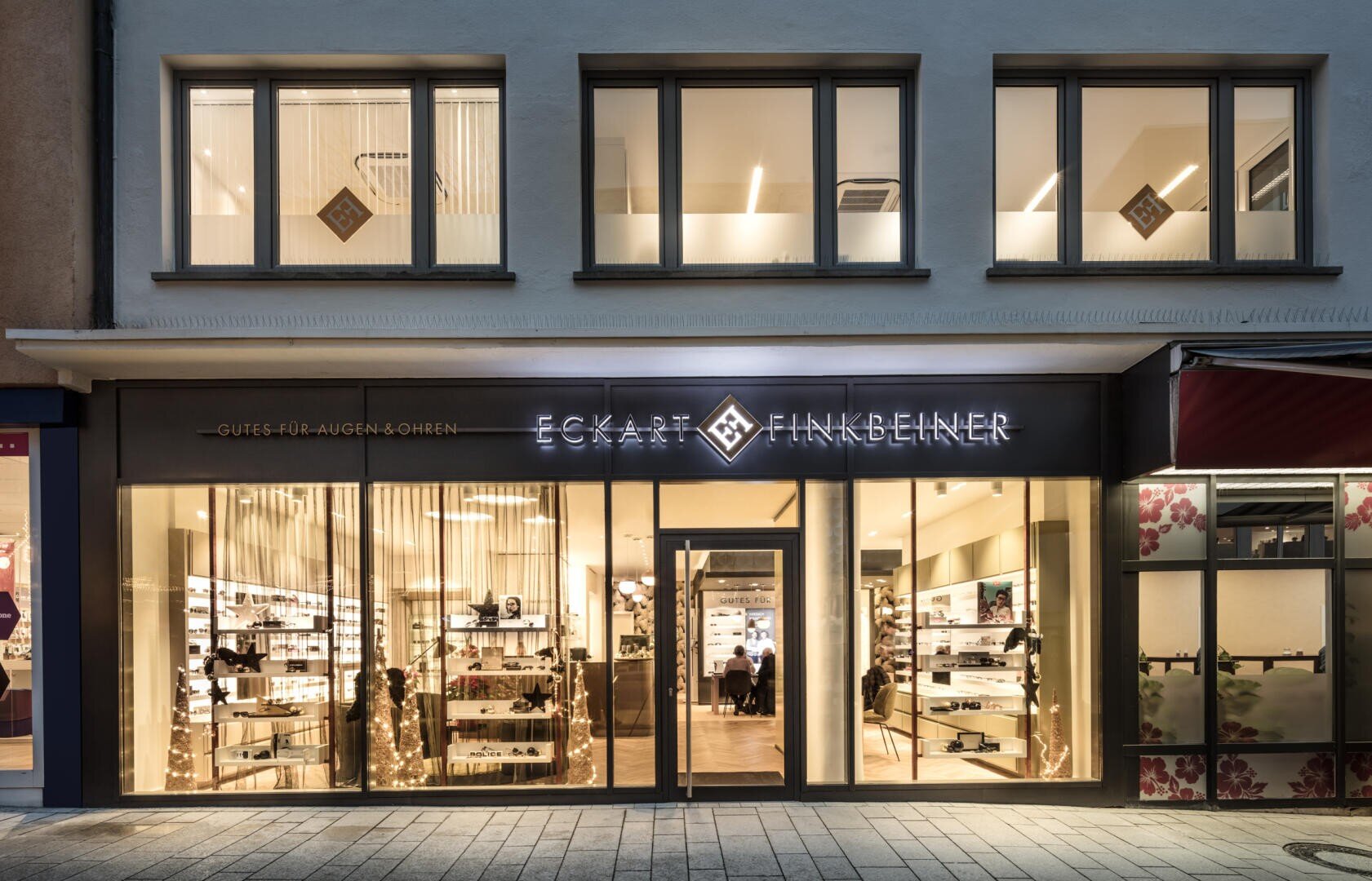 Schaufenster des Optikergeschäfts Eckart Finkbeiner mit großen Schaufenstern, in denen Brillen ausgestellt sind, und einem hellen, modernen Innenraum. Der Name des Geschäfts ist über dem Eingang beleuchtet.